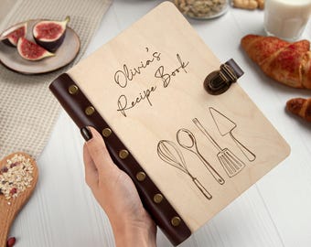 May include: A wooden recipe book with a brown leather spine and metal accents. The cover is engraved with the text "Olivia's Recipe Book" and features a drawing of kitchen utensils.