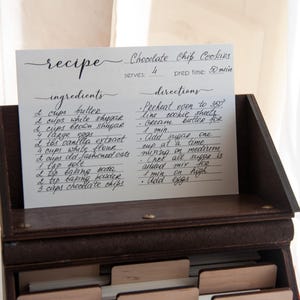 Peut inclure: Une recette manuscrite de cookies aux p&eacute;pites de chocolat est affich&eacute;e dans un coffret &agrave; recettes en bois. La carte &eacute;num&egrave;re les ingr&eacute;dients comme le beurre, le sucre et les p&eacute;pites de chocolat, ainsi que les instructions. La recette est pour 4 personnes et prend 50 minutes.