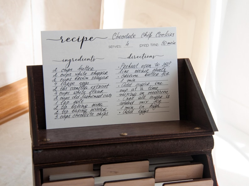 Peut inclure: Une recette manuscrite de cookies aux p&eacute;pites de chocolat est affich&eacute;e dans un coffret &agrave; recettes en bois. La carte &eacute;num&egrave;re les ingr&eacute;dients comme le beurre, le sucre et les p&eacute;pites de chocolat, ainsi que les instructions. La recette est pour 4 personnes et prend 50 minutes.