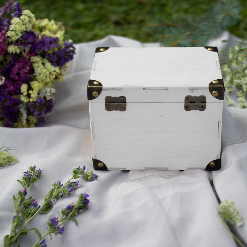 May include: White wooden box with brass hinges and corner accents. The box is closed and sitting on a white fabric surface.