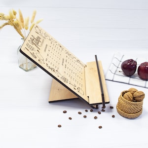May include: A wooden kitchen calendar with a stand. The calendar has a monthly layout with a different kitchen-themed illustration for each month. The calendar is made of light wood and has a dark wood stand.