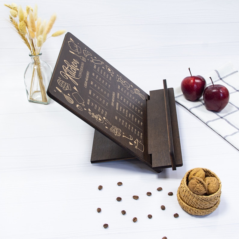 May include: A dark brown wooden kitchen conversion chart stand with a white background. The chart features a variety of kitchen measurements and conversions. The stand is angled and has a small shelf on the bottom.