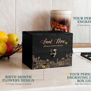 May include: Black wooden box with a floral design and the text "Aunt Nina Recipes & Cooking Secrets". The box has a hinged lid with a metal clasp. The box is on a countertop with a bowl of fruit in the background.