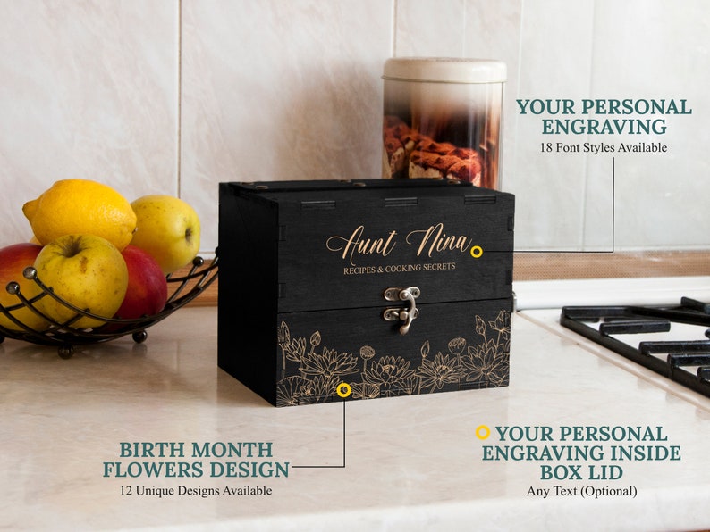 May include: Black wooden box with a floral design and the text "Aunt Nina Recipes & Cooking Secrets". The box has a hinged lid with a metal clasp. The box is on a countertop with a bowl of fruit in the background.