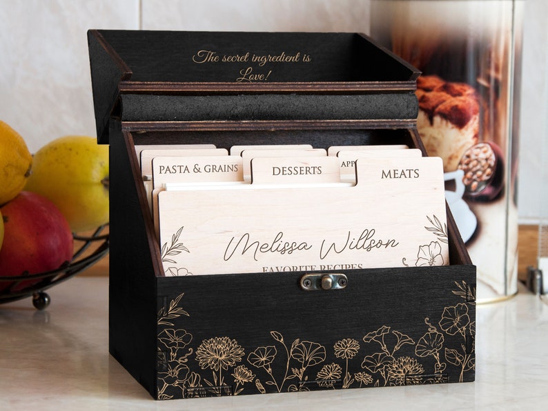 May include: Black wooden recipe box with a floral design and the text "The secret ingredient is Love!" on the top. The box has dividers for different recipe categories, including "Pasta & Grains", "Desserts", "Appetizers", and "Meats". The front of the box has the name "Melissa Willson" and the words "Favorite Recipes".