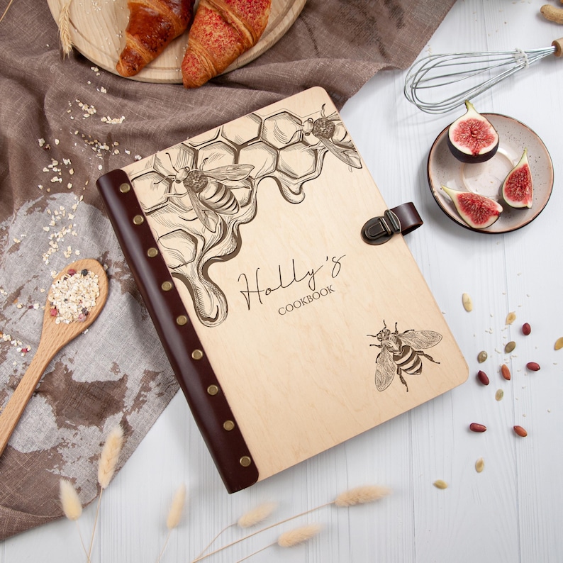 May include: A wooden cookbook with a brown leather cover and a beehive design. The cover is engraved with the text "Holly's Cookbook" and features illustrations of bees and honeycombs.