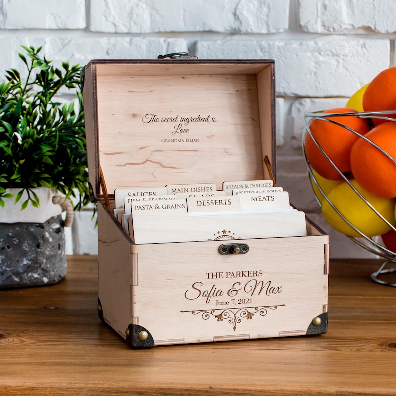 May include: Wooden recipe box with an open lid, featuring the text "The secret ingredient is Love" and "Grandma Lilian." The box is personalized with "Sofia & Max" and "June 7, 2021." It contains labeled dividers for recipes.