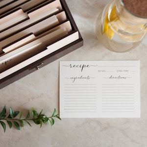 May include: A white recipe card with black text that reads "recipe", "ingredients", "serves", "prep time", and "directions". The card is on a light brown surface with a wooden recipe box, a glass pitcher of water, and a green plant.
