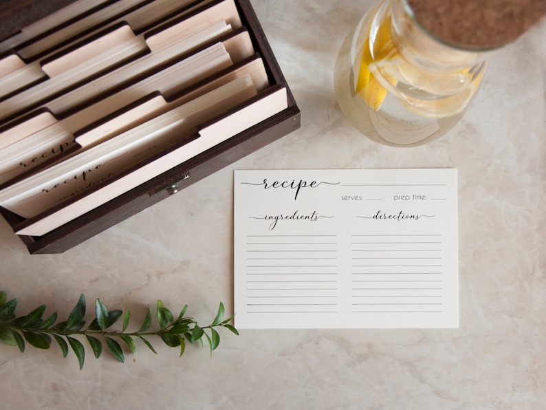May include: A white recipe card with black text that reads "recipe", "ingredients", "serves", "prep time", and "directions". The card is on a light brown surface with a wooden recipe box, a glass pitcher of water, and a green plant.