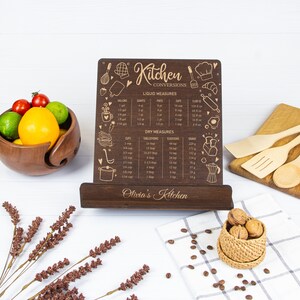 May include: A wooden kitchen conversion chart with a stand. The chart has a gold engraved design with various kitchen measurements for liquid and dry ingredients. The chart is personalized with the name "Olivia's Kitchen".