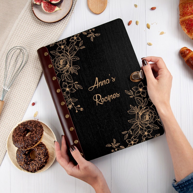 Puede incluir: Libro de recetas negro con un dise&ntilde;o floral dorado y el texto "Anna's Recipes" en la portada.