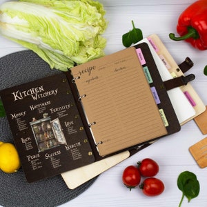 Pu&ograve; includere: Un ricettario rilegato in pelle marrone con una copertina in legno nero che presenta un'illustrazione vintage di un armadio da cucina con barattoli di erbe e spezie. La copertina include anche il testo "Kitchen Witchery" e un elenco di diversi tipi di incantesimi, come "Money", "Happiness", "Fertility", "Love", "Luck", "Peace", "Success" e "Health". Il libro ha un sistema di rilegatura ad anelli e include schede per diverse categorie di ricette, come "Breakfast", "Salads", "Vegetarian", "Drinks", "Side Dishes" e "Miscellaneous".