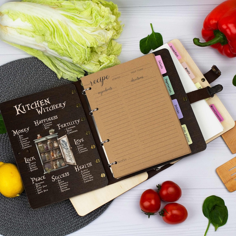 Pu&ograve; includere: Un ricettario rilegato in pelle marrone con una copertina in legno nero che presenta un'illustrazione vintage di un armadio da cucina con barattoli di erbe e spezie. La copertina include anche il testo "Kitchen Witchery" e un elenco di diversi tipi di incantesimi, come "Money", "Happiness", "Fertility", "Love", "Luck", "Peace", "Success" e "Health". Il libro ha un sistema di rilegatura ad anelli e include schede per diverse categorie di ricette, come "Breakfast", "Salads", "Vegetarian", "Drinks", "Side Dishes" e "Miscellaneous".