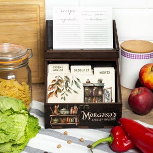 Peut inclure: Une bo&icirc;te &agrave; recettes en bois brun fonc&eacute; avec le texte "Morgana's Spells & Recipes" sur le devant. La bo&icirc;te contient des fiches de recettes et des s&eacute;parateurs &eacute;tiquet&eacute;s "Pasta & Grains", "Desserts" et "Meats". Un bocal de p&acirc;tes, des poivrons rouges et des pommes sont en arri&egrave;re-plan.