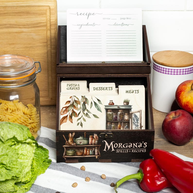 Peut inclure: Une bo&icirc;te &agrave; recettes en bois brun fonc&eacute; avec le texte "Morgana's Spells & Recipes" sur le devant. La bo&icirc;te contient des fiches de recettes et des s&eacute;parateurs &eacute;tiquet&eacute;s "Pasta & Grains", "Desserts" et "Meats". Un bocal de p&acirc;tes, des poivrons rouges et des pommes sont en arri&egrave;re-plan.