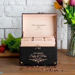 May include: A black wooden recipe box with gold accents, open to reveal recipe dividers. The box is personalized with the names "Sofia & Max" and the date "June 7, 2021." The inside lid reads "The secret ingredient is Love, Grandma Lilian."