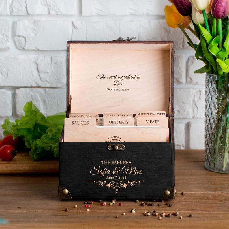 May include: A black wooden recipe box with gold accents, open to reveal recipe dividers. The box is personalized with the names "Sofia & Max" and the date "June 7, 2021." The inside lid reads "The secret ingredient is Love, Grandma Lilian."