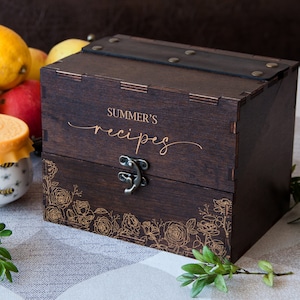 May include: A dark brown wooden recipe box with a leather strap and a brass latch. The box is engraved with the words "SUMMER'S recipes" and a floral design.