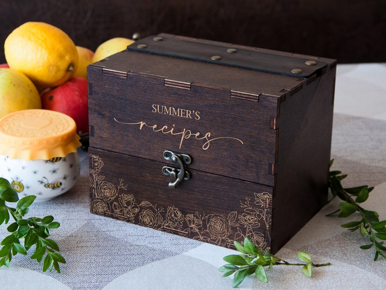 May include: A dark brown wooden recipe box with a leather strap and a brass latch. The box is engraved with the words "SUMMER'S recipes" and a floral design.
