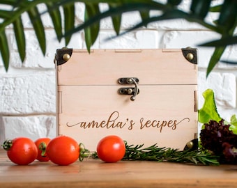 May include: A light wooden recipe box with the words "amelia's recipes" in cursive script. The box has a metal clasp and corner accents. Red cherry tomatoes and greenery are arranged in front of the box.