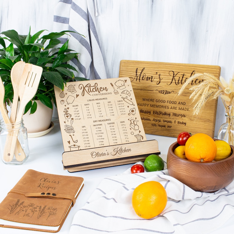 May include: A wooden kitchen stand with a recipe conversion chart and a personalized inscription that reads "Olivia's Kitchen". The stand is next to a brown leather recipe book with the inscription "Olivia's Recipes". A wooden cutting board with the inscription "Mom's Kitchen" is in the background. The cutting board has the text "Where good food and happy memories are made. Happy Birthday Hama Ahlia, Azizi, Umar & Baby 5th August 2021".