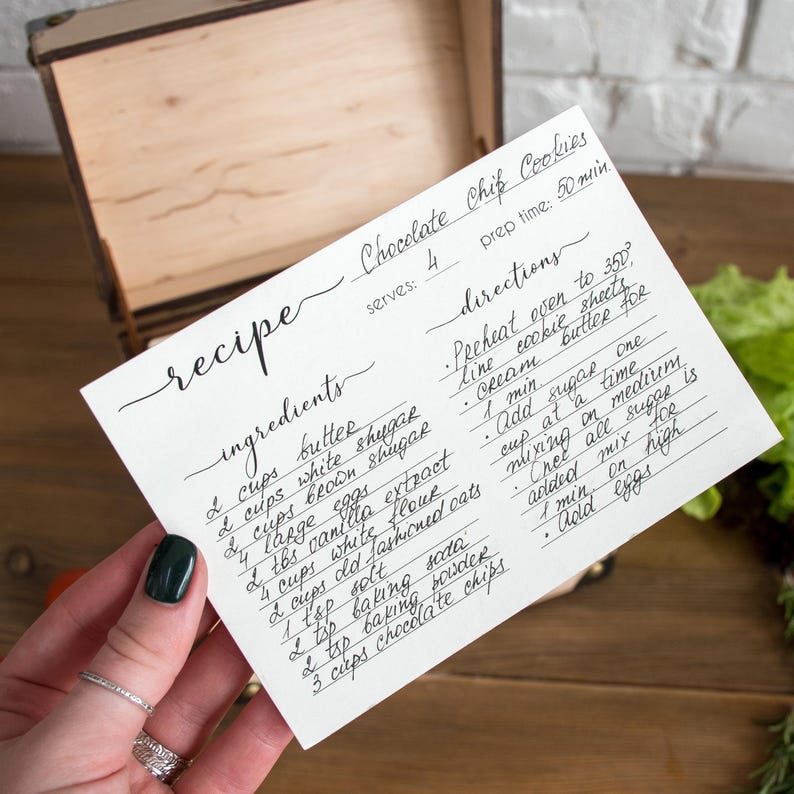 May include: A handwritten recipe card for chocolate chip cookies, held in a hand. The card lists ingredients like butter, sugar, and chocolate chips, and directions for baking. A wooden box is in the background. The prep time is 50 minutes.