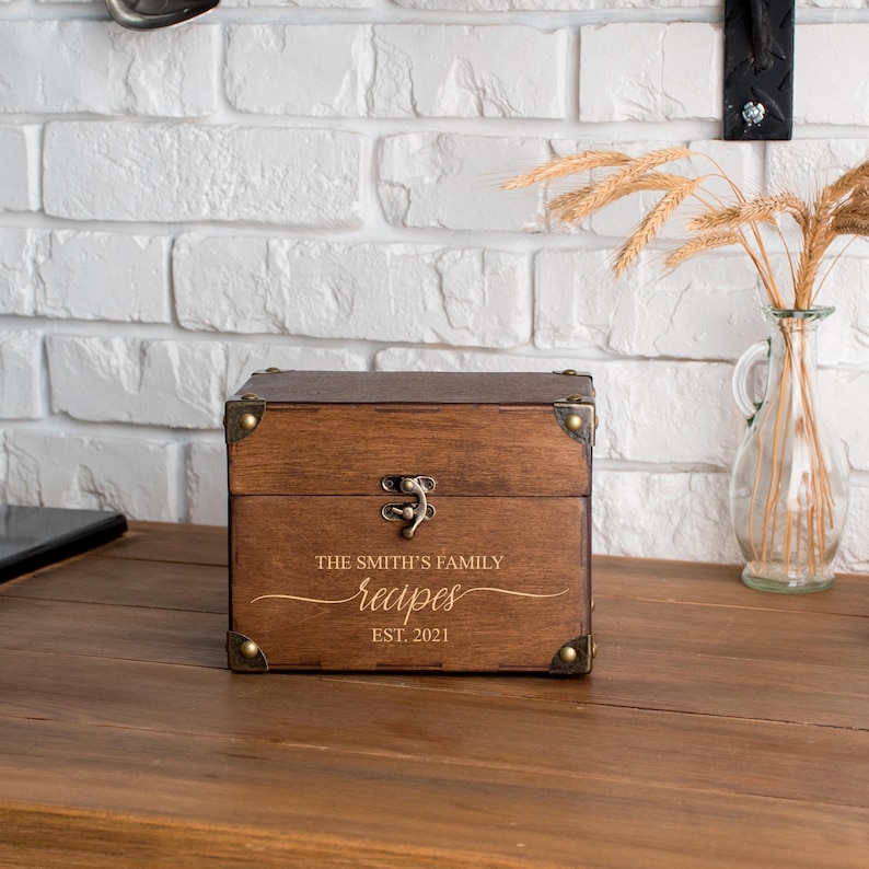 May include: A wooden recipe box with brass corner details and a clasp. The box is engraved with "THE SMITH'S FAMILY recipes EST. 2021". The box is on a wooden surface. The box is a dark brown colour.