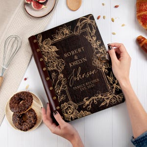 Puede incluir: Un libro de recetas de madera marr&oacute;n oscuro con un dise&ntilde;o floral dorado y el texto "Robert & Joselyn Johnson Recetas Familiares Est. 2025".