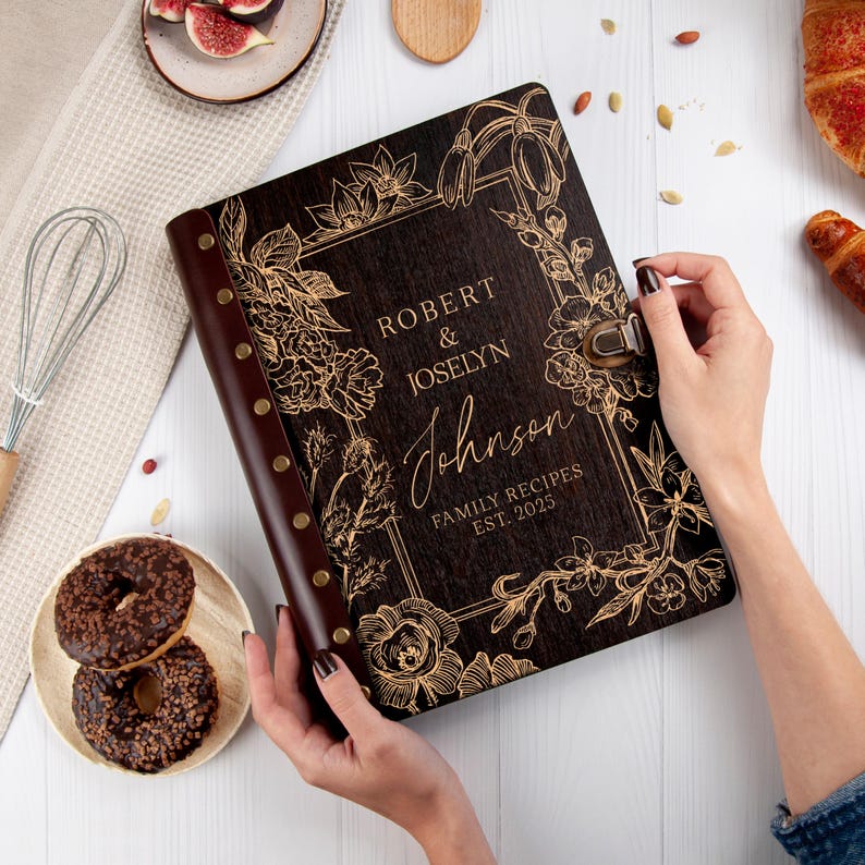 Puede incluir: Un libro de recetas de madera marr&oacute;n oscuro con un dise&ntilde;o floral dorado y el texto "Robert & Joselyn Johnson Recetas Familiares Est. 2025".