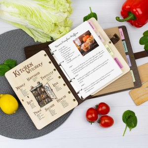 Pu&ograve; includere: Un raccoglitore di ricette rilegato in pelle marrone con una copertina in legno con un tema di stregoneria da cucina. La copertina presenta illustrazioni di barattoli di erbe e spezie, e le parole "Kitchen Witchery" in un carattere decorativo. Il raccoglitore &egrave; aperto a una pagina con una ricetta per i muffin di zucca di Nana.