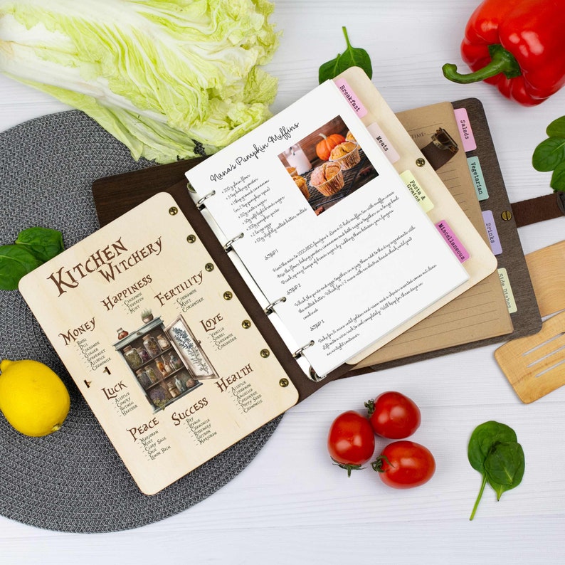 Pu&ograve; includere: Un raccoglitore di ricette rilegato in pelle marrone con una copertina in legno con un tema di stregoneria da cucina. La copertina presenta illustrazioni di barattoli di erbe e spezie, e le parole "Kitchen Witchery" in un carattere decorativo. Il raccoglitore &egrave; aperto a una pagina con una ricetta per i muffin di zucca di Nana.