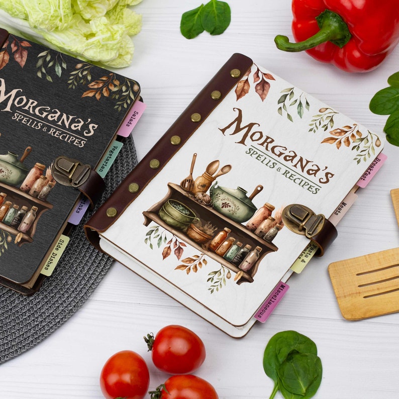 Pu&ograve; includere: Due libri di ricette in bianco e nero con il titolo "Morgana's Spells & Recipes" e un'illustrazione colorata di uno scaffale da cucina con varie bottiglie e barattoli. I libri hanno schede per diverse categorie, tra cui "Colazione", "Carne", "Pasta e piatti principali", "Insalate", "Vegetariano", "Contorni" e "Varie".
