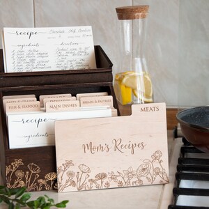 May include: A wooden recipe box with a floral design and the text "Mom's Recipes". The box has dividers for different categories of recipes, such as "Fish & Seafood", "Main Dishes", and "Meats".