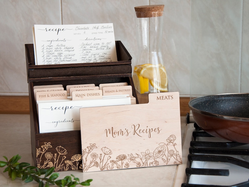 May include: A wooden recipe box with a floral design and the text "Mom's Recipes". The box has dividers for different categories of recipes, such as "Fish & Seafood", "Main Dishes", and "Meats".