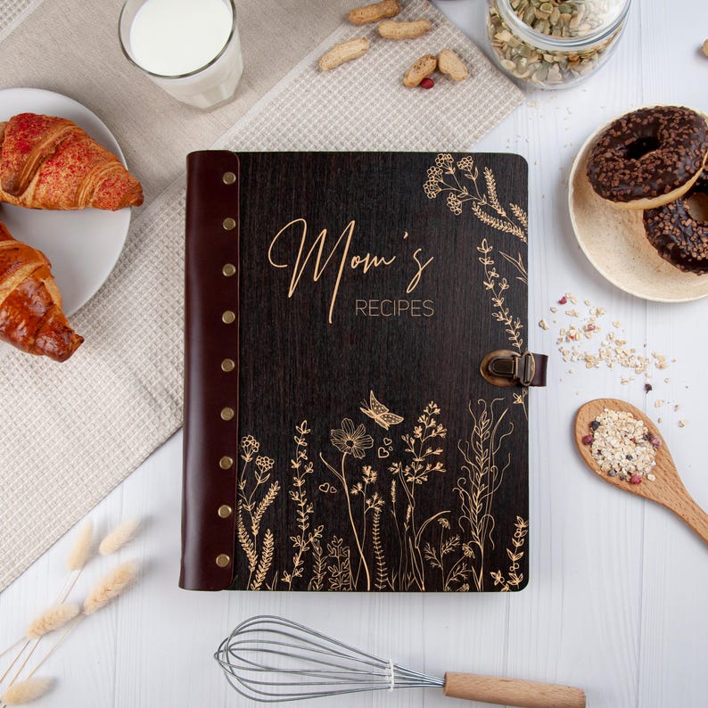 May include: A dark brown wooden recipe book with a leather spine and gold accents. The cover features a floral design and the text "Mom's Recipes".