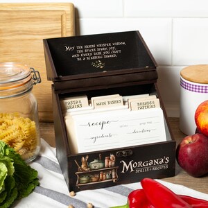Peut inclure: Un coffret &agrave; recettes en bois brun fonc&eacute; avec le texte "May the herbs whisper wisdom..." sur le couvercle. La bo&icirc;te contient des fiches de recettes et des s&eacute;parateurs &eacute;tiquet&eacute;s "Pasta & Grains", "Main Dishes" et "Breads & Pastries". La bo&icirc;te est d&eacute;cor&eacute;e d'illustrations et du texte "Morgana's Spells & Recipes."