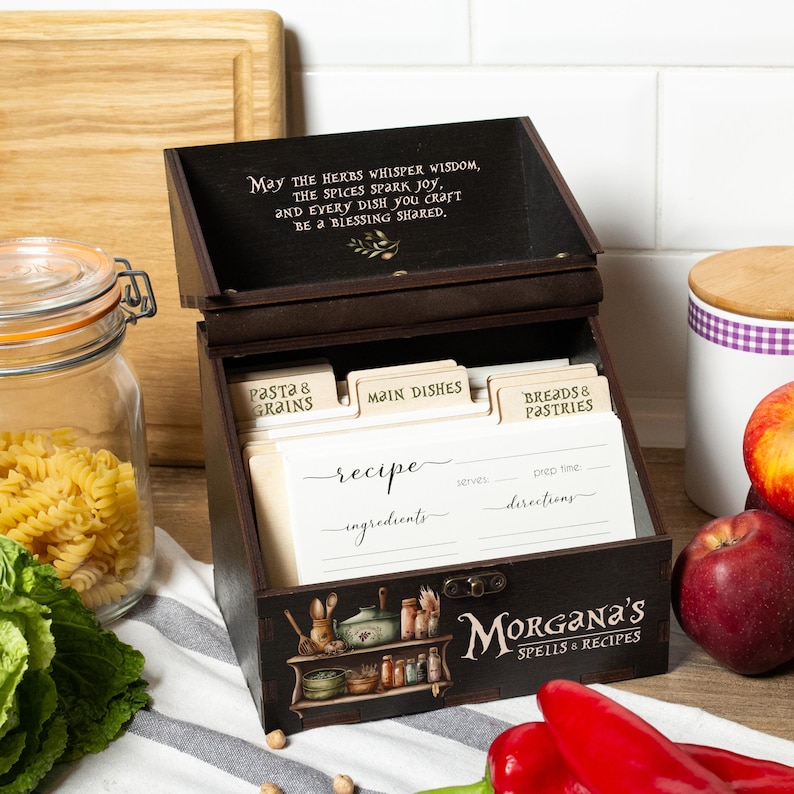 Peut inclure: Un coffret &agrave; recettes en bois brun fonc&eacute; avec le texte "May the herbs whisper wisdom..." sur le couvercle. La bo&icirc;te contient des fiches de recettes et des s&eacute;parateurs &eacute;tiquet&eacute;s "Pasta & Grains", "Main Dishes" et "Breads & Pastries". La bo&icirc;te est d&eacute;cor&eacute;e d'illustrations et du texte "Morgana's Spells & Recipes."