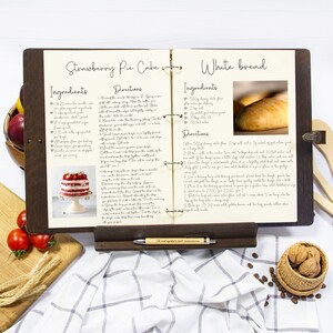 May include: Open recipe book with two pages showing recipes for Strawberry Pie Cake and White Bread. The Strawberry Pie Cake recipe includes ingredients like sponge cake, strawberries, and whipped cream. The White Bread recipe includes ingredients like flour, yeast, and water.