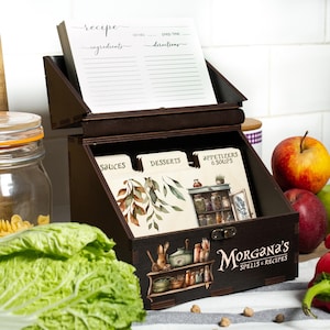 Peut inclure: Un coffret &agrave; recettes en bois brun fonc&eacute; avec un couvercle &agrave; charni&egrave;re, contenant des fiches de recettes et des s&eacute;parateurs &eacute;tiquet&eacute;s "Sauces", "Desserts" et "Entr&eacute;es & Soupes". La bo&icirc;te est d&eacute;cor&eacute;e d'illustrations et du texte "Morgana's Spells & Recipes."
