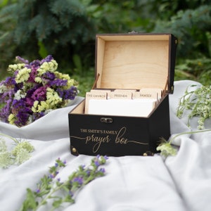 May include: A black wooden box with a gold inscription that reads "The Smith's Family Prayer Box". The box has a hinged lid and is open to reveal a set of dividers labeled "The Church", "Friends", and "Family".
