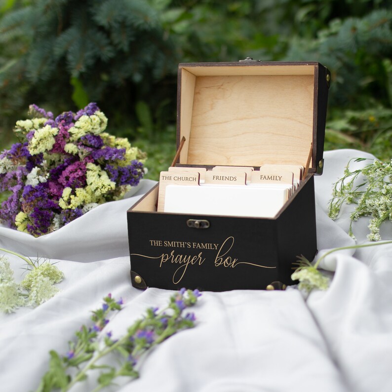 May include: A black wooden box with a gold inscription that reads "The Smith's Family Prayer Box". The box has a hinged lid and is open to reveal a set of dividers labeled "The Church", "Friends", and "Family".