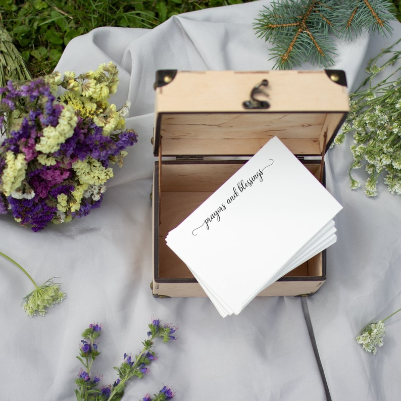May include: A wooden box with a lid open, revealing a stack of white cards with the text "Prayers and Blessings" written in black calligraphy. The box is sitting on a white fabric surface with dried flowers and greenery around it.