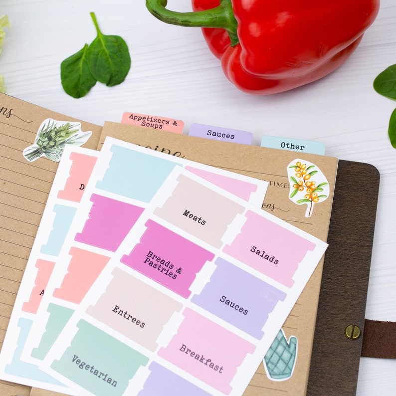Pu&ograve; includere: Un set di divisori per ricette colorati con schede per diverse categorie, tra cui antipasti, zuppe, carni, pane e pasticceria, insalate, salse, piatti principali, colazione e vegetariani. I divisori sono su un taccuino marrone con un peperone rosso sullo sfondo.