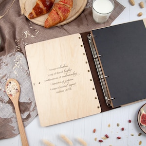 May include: A wooden photo album with a black leather cover and a ring binder. The front cover is engraved with the words "2 cups of kindness, 1 cup of shared laughter, 3 tablespoons of understanding, 1 teaspoon of patience, 4 cups of mutual respect, endless LOVE".