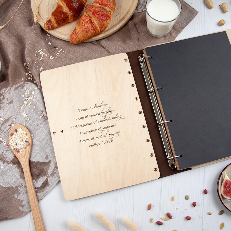 May include: A wooden photo album with a black leather cover and a ring binder. The front cover is engraved with the words "2 cups of kindness, 1 cup of shared laughter, 3 tablespoons of understanding, 1 teaspoon of patience, 4 cups of mutual respect, endless LOVE".