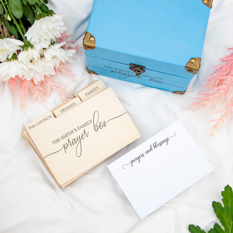 May include: A wooden prayer box with dividers for different categories, such as "The Church", "Friends", and "Family". The box is labeled "The Smith's Family Prayer Box". A white card with the text "prayers and blessings" is also shown.