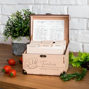 May include: A wooden recipe box, engraved with "Tamling's Favorite Recipes" and a chef's hat. Inside, there are recipe cards and a handwritten chocolate chip cookie recipe. Index cards are labelled for various food categories, such as "Sauces" and "Salads".