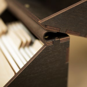 May include: Close-up of a wooden box with a leather hinge. The box is made of dark wood and has a light wood interior. The hinge is made of leather and has a metal rivet.