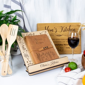 May include: A wooden recipe stand with a brown leather recipe book that says "Olivia's Recipes" on the cover. The stand is engraved with the words "Olivia's Kitchen". There is a wooden cutting board behind the stand that says "Mom's Kitchen" with a birthday message.