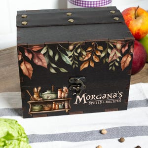 Peut inclure: Une bo&icirc;te en bois noire avec un couvercle &agrave; charni&egrave;re et un fermoir en m&eacute;tal. La bo&icirc;te est d&eacute;cor&eacute;e d'un motif botanique de feuilles dans des tons de marron, vert et orange. Le texte "Morgana's Spells & Recipes" est affich&eacute;.